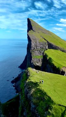 Ziyaretçiler yemyeşil araziyi keşfederken Faroe Adaları 'ndaki okyanus üzerinde nefes kesen uçurumlar belirgin bir şekilde yükseliyor. Mylingur uçurumunda yürüyüş yapan bir çift.