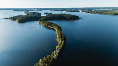 Finlandiya, Punkaharju 'daki yemyeşil ve dingin suların ortasındaki dolambaçlı esker yolunun nefes kesici manzarası. Sahne, Finlandiya 'nın gündüz doğa ve manzarasının güzelliğini yakalar..