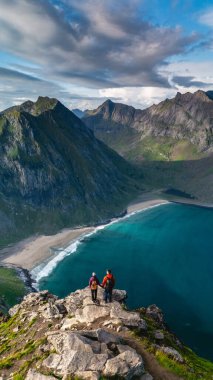 Yürüyüşçüler, Norveç 'in Lofoten kentindeki Kvalvika plaj patikasındaki Ryten zirvesinden çarpıcı manzaraların keyfini çıkarıyorlar. Sakin turkuaz sular ve engebeli dağlar macera için mükemmel bir zemin oluşturur..