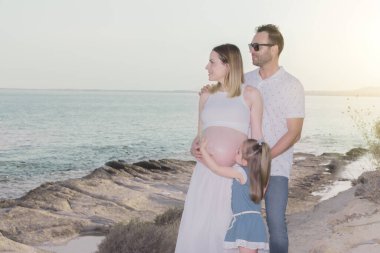 Genç bir aile, hamile bir anneyle deniz kenarında geziniyor. Küçük kız annesinin karnını öpüyor.