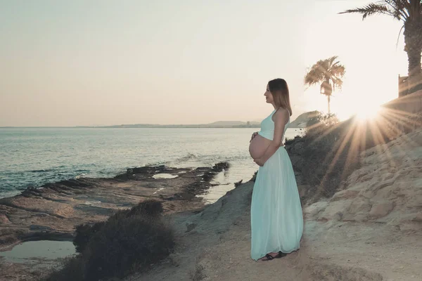 Genç hamile bir kadın plajda karnına sevgiyle dokunuyor ve gün batımında sakin denizde dinleniyor.