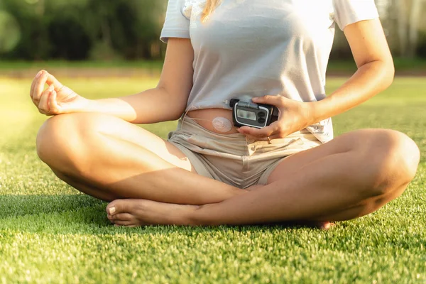 Diyabet hastası bir kadın yoga yapıyor. Kız elinde bir insülin pompası tutuyor. Parktaki çimlerin üzerinde bir kadın oturuyor. Diyabetli sağlıklı bir yaşam tarzı