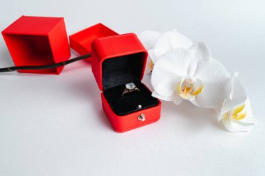 Nişan yüzüğü kırmızı hediye kutusunda, beyaz arka planda. Evlen benimle. Lüks aksesuar. Mesaj için yer. Tropik orkide çiçekleri.