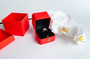 Nişan yüzüğü kırmızı hediye kutusunda, beyaz arka planda. Evlen benimle. Lüks aksesuar. Mesaj için yer. Tropik orkide çiçekleri.