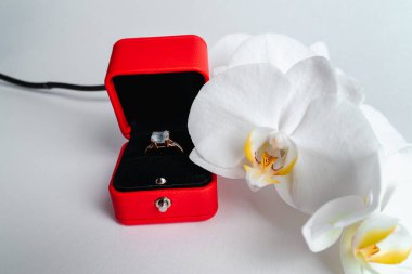 Nişan yüzüğü kırmızı hediye kutusunda, beyaz arka planda. Evlen benimle. Lüks aksesuar. Mesaj için yer. Tropik orkide çiçekleri.