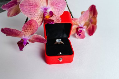 Nişan yüzüğü kırmızı hediye kutusunda, beyaz arka planda. Evlen benimle. Lüks aksesuar. Mesaj için yer. Tropik orkide çiçekleri.