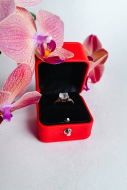 Nişan yüzüğü kırmızı hediye kutusunda, beyaz arka planda. Evlen benimle. Lüks aksesuar. Mesaj için yer. Tropik orkide çiçekleri.