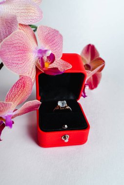 Nişan yüzüğü kırmızı hediye kutusunda, beyaz arka planda. Evlen benimle. Lüks aksesuar. Mesaj için yer. Tropik orkide çiçekleri.