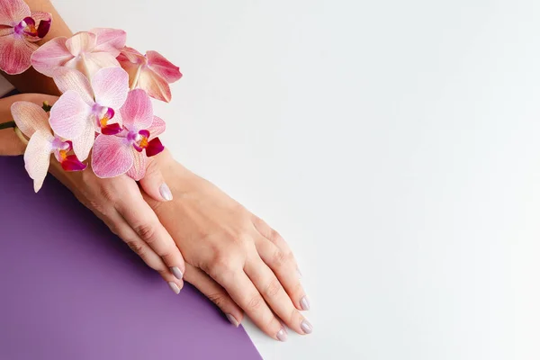 Young woman\'s hands close-up. Stylish trendy manicure on white and purple background. Tropical orchid flowers. Place for text. Advertising of manicure and beauty salon. Relaxation spa service