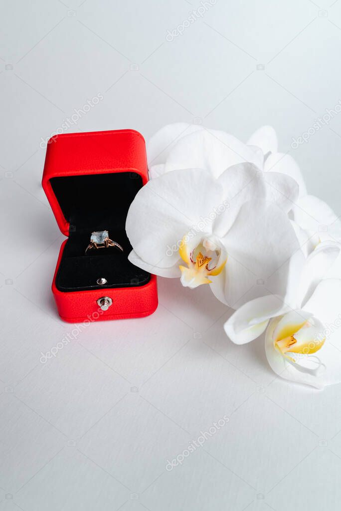 Engagement ring in red gift box on white background.  Marry me. Luxury accessory. Place for text. Tropical orchid flowers.