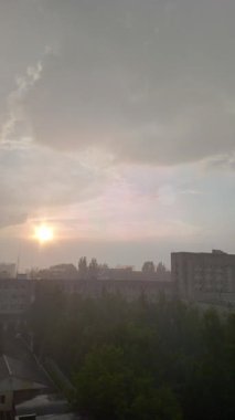 Şehir manzarasında yağmurlu günbatımını yakalayan dikey video, bulutları aşan güneş ışığı ve şehir akşam manzarasında yağış ile nadir görülen hava fenomeni