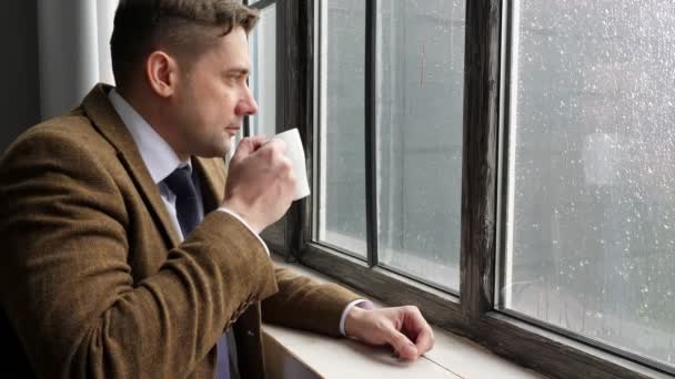 Молодий чоловік у костюмі п'є чай, стоячи біля вікна під час дощу . — стокове відео