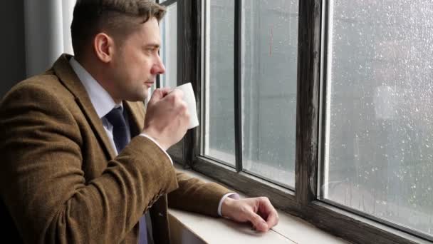 Un joven con traje bebe té mientras está de pie junto a la ventana bajo la lluvia . — Vídeos de Stock