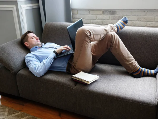 Un hombre yace en su casa en el sofá y trabaja en una computadora portátil. Concepto de trabajo remoto . — Foto de Stock