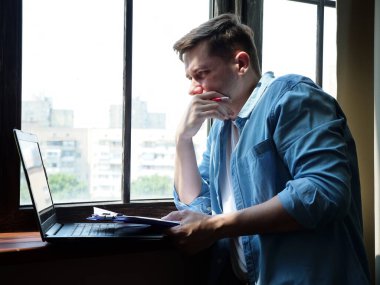 Penceredeki genç çekici adam dizüstü bilgisayarla çalışıyor. Serbest ve uzak çalışma.