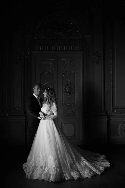Schwarz-Weiß-Fotografie. Luxus-Hochzeitspaar verliebt. schöne Braut im weißen Kleid mit Brautstrauß und schöner Bräutigam im schwarzen Anzug, der im barocken Interieur steht und sich umarmt — Stockfoto