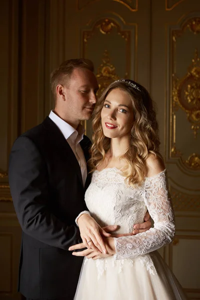 Pareja de bodas de lujo enamorada. Hermosa novia en vestido blanco con ramo de novias y novio guapo en traje negro de pie en el interior barroco y abrazándose — Foto de Stock