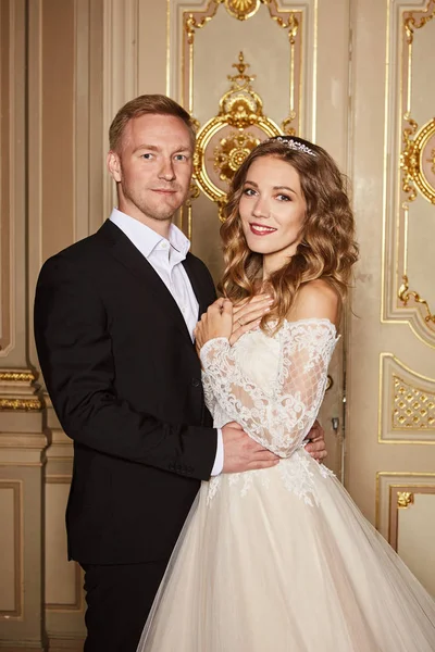 Luxury wedding couple in love. Beautiful bride in white dress with brides bouquet and handsome groom in black suit standing in baroque interior and embracing each other