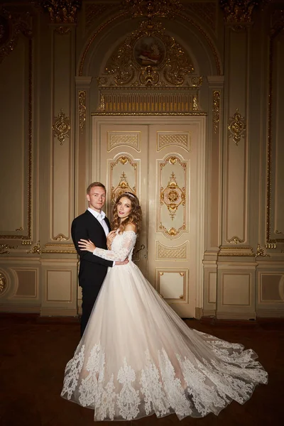 Casal de luxo apaixonado. Bela noiva em vestido branco com buquê de noivas e belo noivo em terno preto em pé no interior barroco e abraçando uns aos outros — Fotografia de Stock