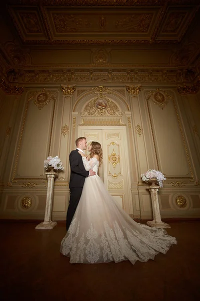 Pareja de bodas de lujo enamorada. Hermosa novia en vestido blanco con ramo de novias y novio guapo en traje negro de pie en el interior barroco y abrazándose — Foto de Stock