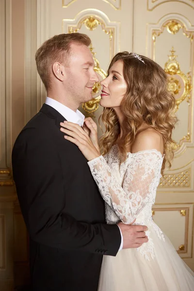 Luxus-Hochzeitspaar verliebt. schöne Braut im weißen Kleid mit Brautstrauß und schöner Bräutigam im schwarzen Anzug, der im barocken Interieur steht und sich umarmt — Stockfoto