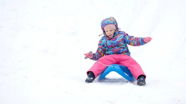 在雪地覆盖的雪橇小径上，一个快乐的女孩骑着雪橇下山，在一个阳光明媚的冬季山景中 — 图库照片