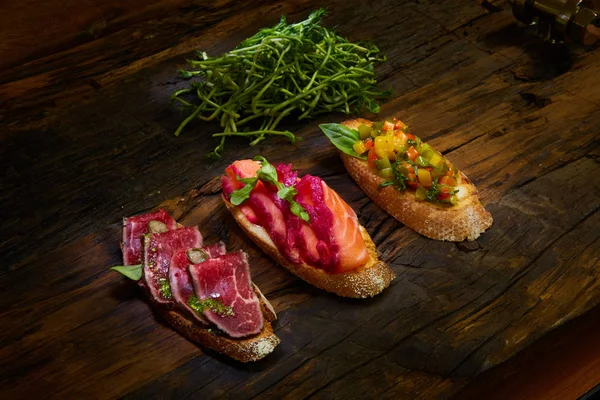 Bruschetta assortita con arrosto di manzo, verdure e salmone leggermente salato con foglie verdi su fondo di legno . — Foto Stock