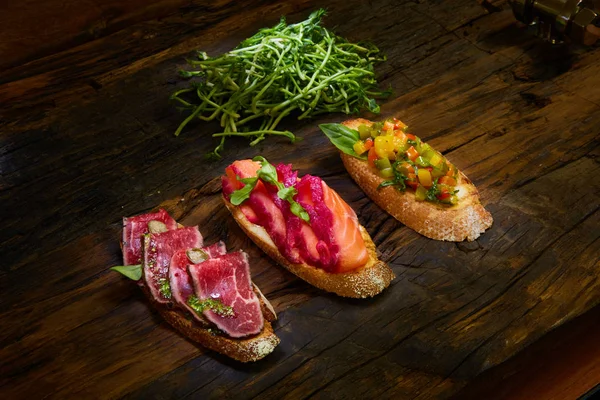 Bruschetta assortita con arrosto di manzo, verdure e salmone leggermente salato con foglie verdi su fondo di legno . — Foto Stock