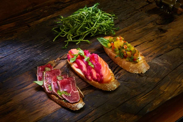 Bruschetta assortita con arrosto di manzo, verdure e salmone leggermente salato con foglie verdi su fondo di legno . — Foto Stock