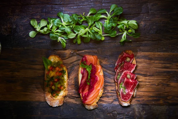 Assorted bruschetta with roast beef, vegetables and lightly salted salmon with greens leaves on wooden background. — Stock Photo, Image