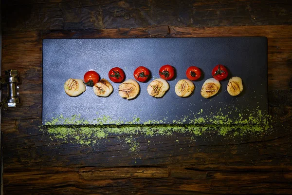 Fried scallops with tomatoes on a black plate. Shallow dof. — Stock Photo, Image