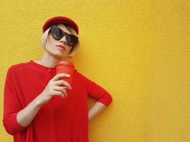 Uzun kırmızı kazak ve kırmızı kağıt Kupası duran sarı zemin üzerine kırmızı şapkalı kadın model. Kazaklı güzel beyaz kadının açık fotoğraf önünde Cafe kahve içer
