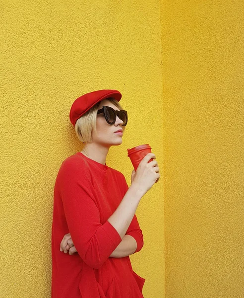 Vrouwelijke model in lange rode trui en rode hoed met rode papier beker staande op gele achtergrond. Buiten foto van mooie Kaukasische vrouw in trui drinkt koffie tegenover café — Stockfoto