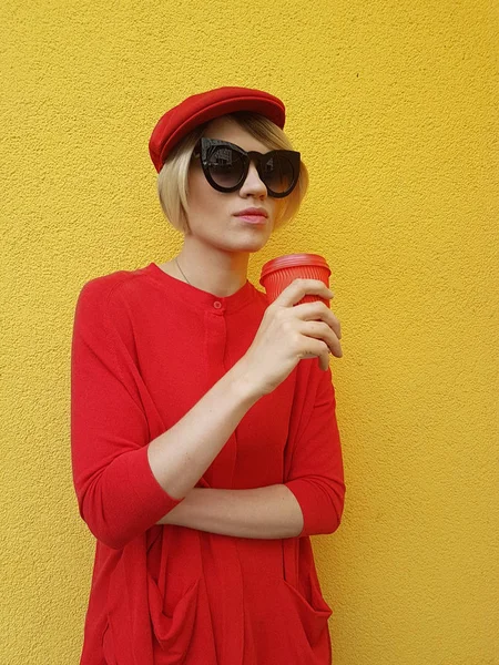 Modèle féminin en pull long rouge et chapeau rouge avec tasse en papier rouge debout sur fond jaune. Photo extérieure de belle femme caucasienne en pull boit du café devant le café — Photo