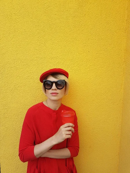 Modelo feminino em camisola vermelha longa e chapéu vermelho com copo de papel vermelho em pé sobre fundo amarelo. Foto ao ar livre da bela mulher caucasiana em suéter bebe café na frente do café — Fotografia de Stock