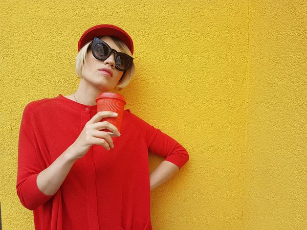 Modelo feminino em camisola vermelha longa e chapéu vermelho com copo de papel vermelho em pé sobre fundo amarelo. Foto ao ar livre da bela mulher caucasiana em suéter bebe café na frente do café — Fotografia de Stock