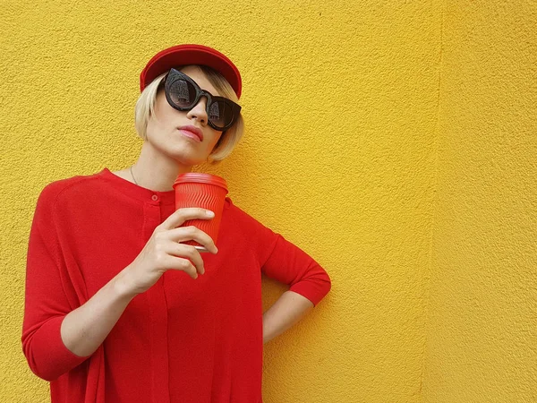 Female model in long red sweater and red hat with red paper cup standing on yellow background. Outdoor photo of beautiful caucasian woman in sweater drinks coffee in front of cafe — Stock Photo, Image