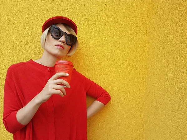 Modèle féminin en pull long rouge et chapeau rouge avec tasse en papier rouge debout sur fond jaune. Photo extérieure de belle femme caucasienne en pull boit du café devant le café — Photo