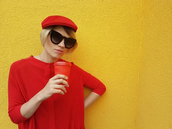 Modelo femenino en jersey rojo largo y sombrero rojo con taza de papel rojo de pie sobre fondo amarillo. Foto al aire libre de la hermosa mujer caucásica en suéter bebe café en frente de la cafetería — Foto de Stock