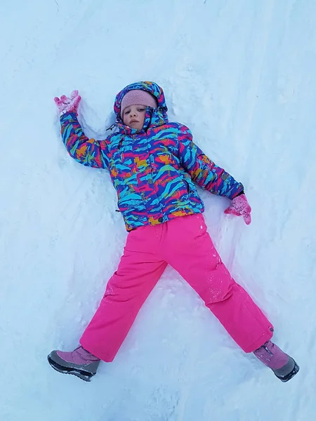 Nettes kleines Mädchen im warmen Skianzug, das draußen Schnee-Engel bastelt. Kinder haben Spaß, wenn sie nach einem Schneesturm im Winter auf Schneewehen liegen. Aktivitäten für Kinder im Freien. Winterurlaub und Urlaub. — Stockfoto