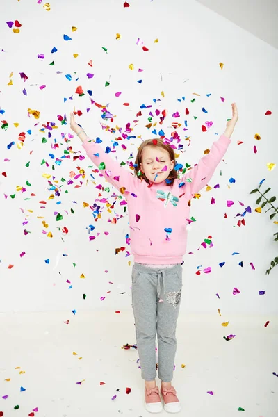 Feliz hermosa niña disfrutando de colorida sorpresa confeti cayendo, posando en la pared del estudio blanco. Chica bonita celebrando su fiesta de cumpleaños, divirtiéndose — Foto de Stock