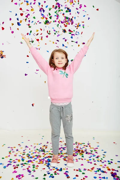 Happy beautiful little girl enjoying colorful confetti surprise falling down, posing on white studio wall. Pretty girl celebrating her birthday party, having fun