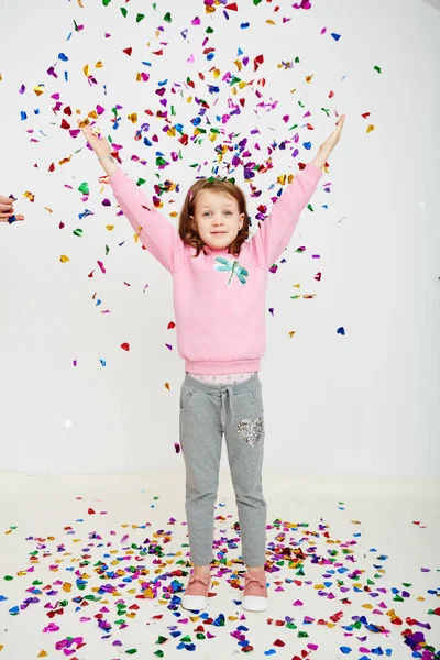 Feliz hermosa niña disfrutando de colorida sorpresa confeti cayendo, posando en la pared del estudio blanco. Chica bonita celebrando su fiesta de cumpleaños, divirtiéndose — Foto de Stock