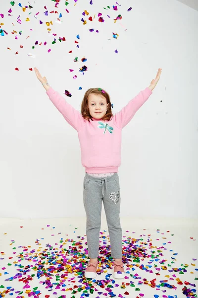Feliz hermosa niña disfrutando de colorida sorpresa confeti cayendo, posando en la pared del estudio blanco. Chica bonita celebrando su fiesta de cumpleaños, divirtiéndose — Foto de Stock