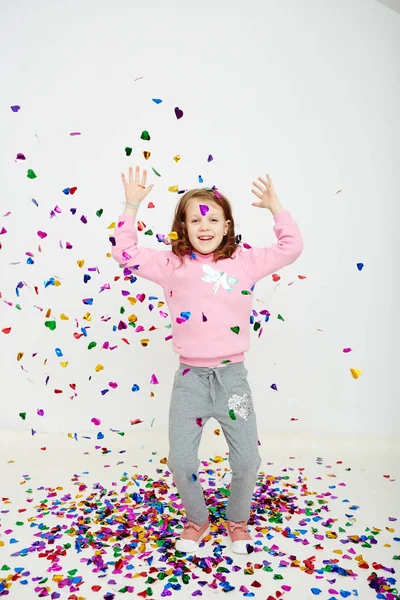 Happy beautiful little girl enjoying colorful confetti surprise falling down, posing on white studio wall. Pretty girl celebrating her birthday party, having fun