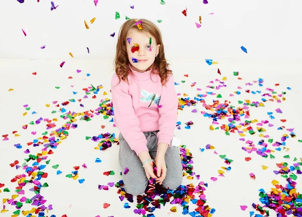 Happy beautiful little girl enjoying colorful confetti surprise falling down, posing on white studio wall. Pretty girl celebrating her birthday party, having fun — Stock Photo, Image