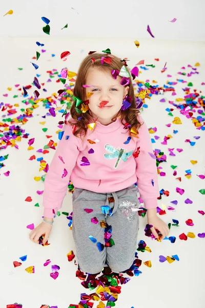 Felice bella bambina godendo sorpresa coriandoli colorati cadere, in posa su parete studio bianco. Bella ragazza che celebra la sua festa di compleanno, divertirsi — Foto Stock