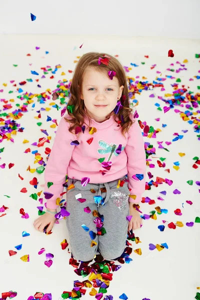 カラフルな紙吹雪驚き落ちて、白いスタジオの壁にポージングを楽しんで幸せな美しい小さな女の子。かわいい女の子の楽しい時を過す彼女の誕生日パーティーを祝う — ストック写真