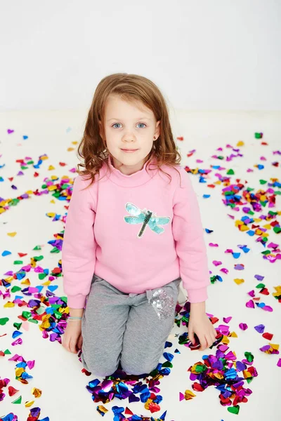 Feliz hermosa niña disfrutando de colorida sorpresa confeti cayendo, posando en la pared del estudio blanco. Chica bonita celebrando su fiesta de cumpleaños, divirtiéndose — Foto de Stock