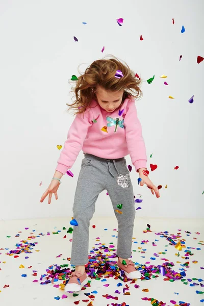 Happy beautiful little girl enjoying colorful confetti surprise falling down, posing on white studio wall. Pretty girl celebrating her birthday party, having fun — Stock Photo, Image